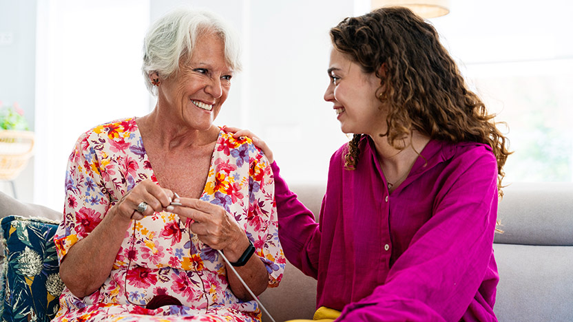 An older woman and a younger woman sit together on a couch, smiling warmly at each other. The older woman wears a floral dress and knits, while the younger woman, in a bright pink blouse, rests her hand on the other’s shoulder.