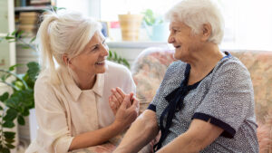 A younger woman with blonde hair smiles and holds hands with an elderly woman with white hair, who is sitting on a sofa. They appear happy and are engaged in a warm conversation in a cozy, sunlit room.
