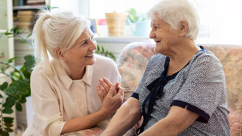 A younger woman with blonde hair smiles and holds hands with an elderly woman with white hair, who is sitting on a sofa. They appear happy and are engaged in a warm conversation in a cozy, sunlit room.