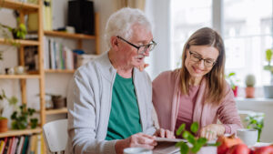 An older man and a younger woman sit together at a table, looking at a tablet and smiling. Bookshelves and plants are visible in the background, creating a cozy, well-lit room.