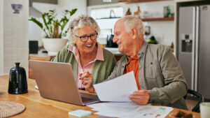 An older couple sits at a kitchen table, smiling as they look at paperwork and a laptop. The scene is bright and homey, with papers, a coffee pot, and plants visible in the background.