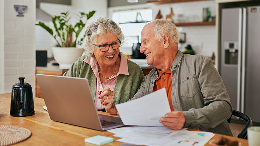 An older couple sits at a kitchen table, smiling as they look at paperwork and a laptop. The scene is bright and homey, with papers, a coffee pot, and plants visible in the background.