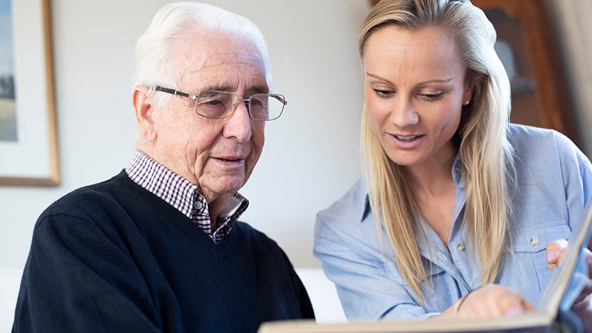 A young woman shows a photo album to an elderly man wearing glasses and a sweater. They are sitting together indoors, looking at the photos and smiling.