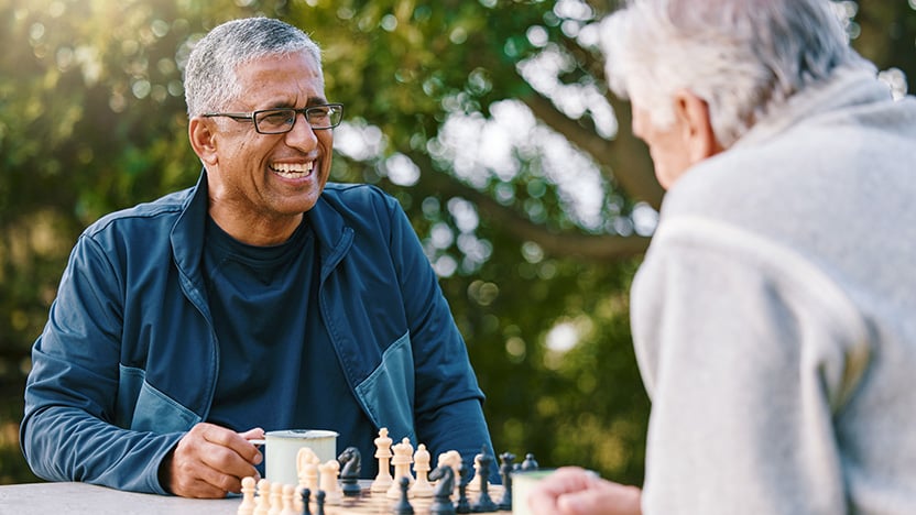 Two older men sit outdoors at a table, smiling and talking while playing chess. One holds a mug, and both appear relaxed and engaged. Trees and greenery are visible in the background.