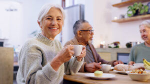 An elderly woman smiles while holding a cup at a table, with two other seniors enjoying food and drinks in a cozy, well-lit kitchen setting.