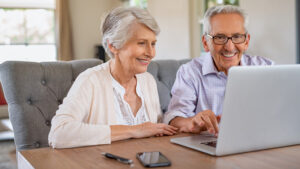 Smiling older couple sitting at a table, looking at a laptop together. A smartphone and pen are on the table in front of them. They appear happy and engaged with what’s on the screen.