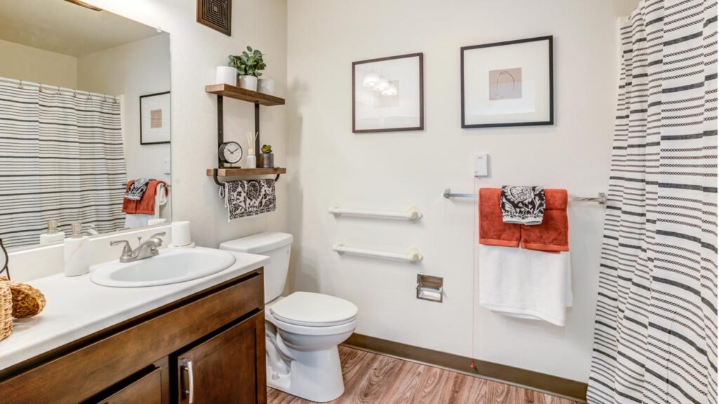 A modern bathroom with a sink, mirror, and wooden cabinet on the left, a toilet in the center, towel racks with red and white towels, wall art, shelves, and a shower with a striped curtain on the right.