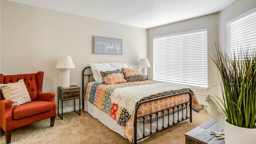 Bright bedroom with a metal bed frame, colorful quilt, two white lamps on bedside tables, an orange armchair, a decorative plant, and a wall sign reading "This is the good life." Large windows bring in natural light.