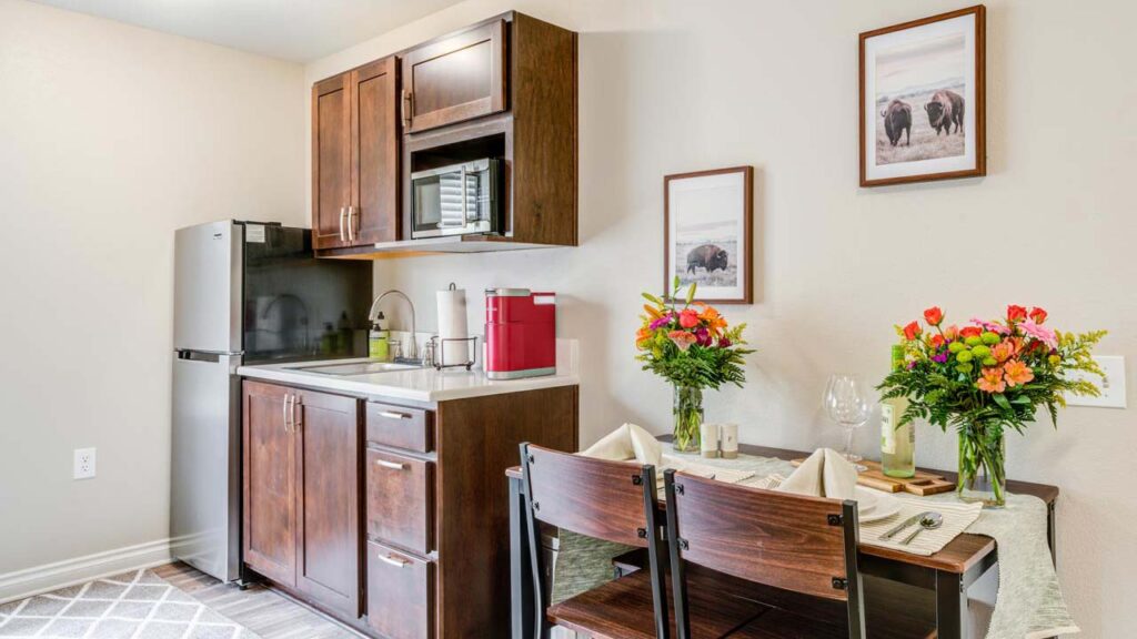 A small kitchen area with dark wood cabinets, a refrigerator, microwave, sink, and coffee maker next to a dining table set for two with flowers and framed animal pictures on the wall.