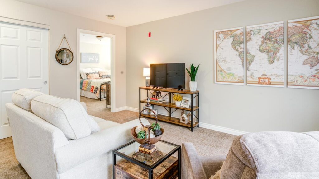 A cozy living room with light-colored furniture, a TV on a black shelf, houseplants, wall art including a world map, and a view into a bedroom with patterned bedding.