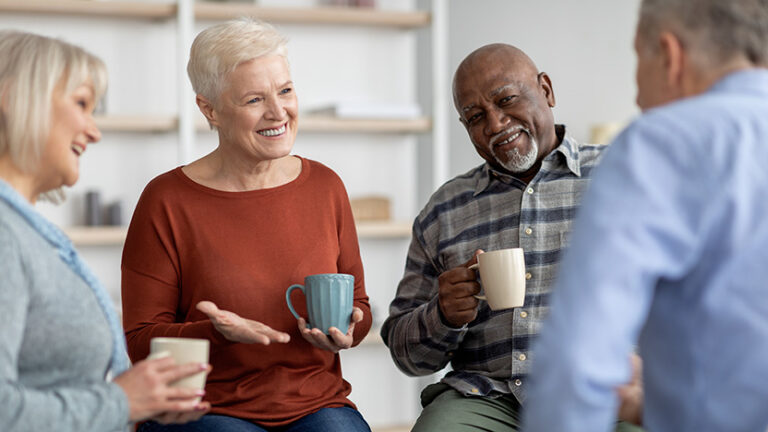 Four older adults sit together, smiling and talking while holding coffee mugs, in a bright, cozy room with shelves in the background.