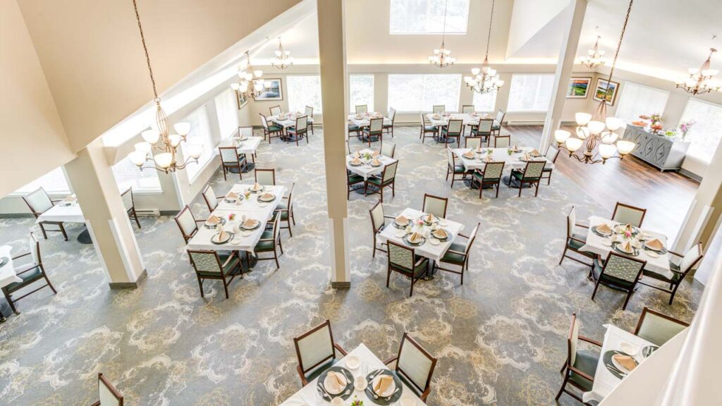 A bright, spacious dining room with several square tables set for meals, each with four chairs, plates, cups, and cutlery. Large windows and elegant chandeliers provide abundant natural and artificial light.
