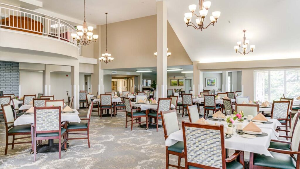 Bright, spacious dining room with neatly set tables, cushioned chairs, elegant chandeliers, and large windows letting in natural light; decorated with flowers and artwork on the walls.