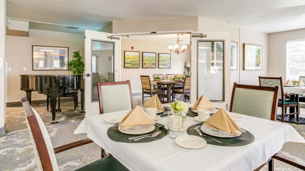 A dining room with a table set for four, tan napkins folded on white plates, and a centerpiece of flowers. In the background, there is a grand piano and framed art on the wall, with bright natural light from windows.