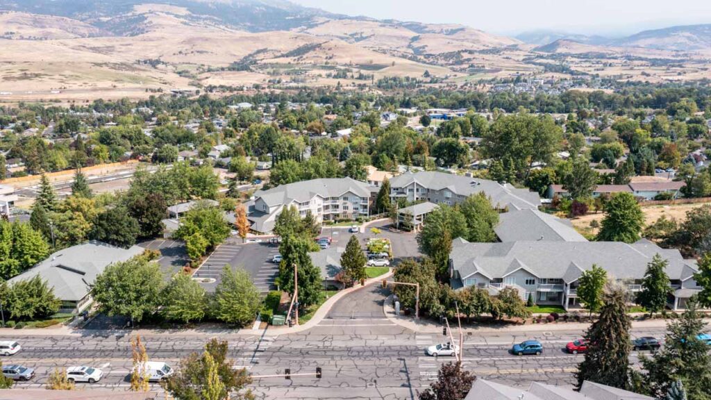 Aerial view of a suburban neighborhood with large buildings surrounded by trees, a main road with cars in front, and rolling hills and mountains in the background under a hazy sky.