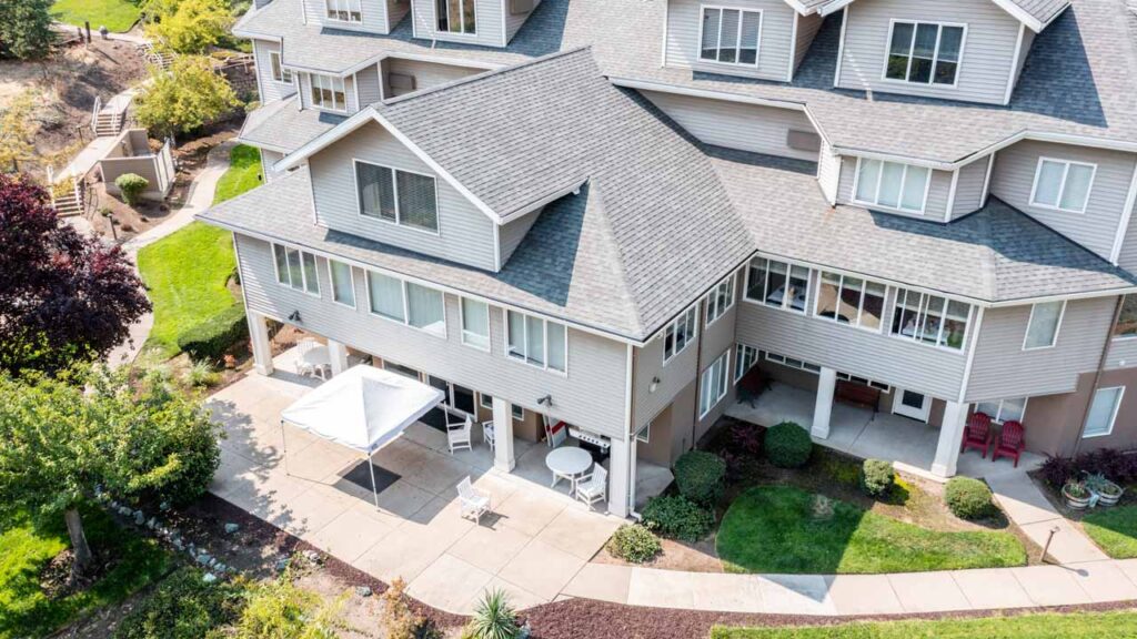 Aerial view of a large, modern, multi-story house with gray siding, white trim, and multiple gabled roofs. The patio below features outdoor seating, a canopy, and is surrounded by green lawns and trees.