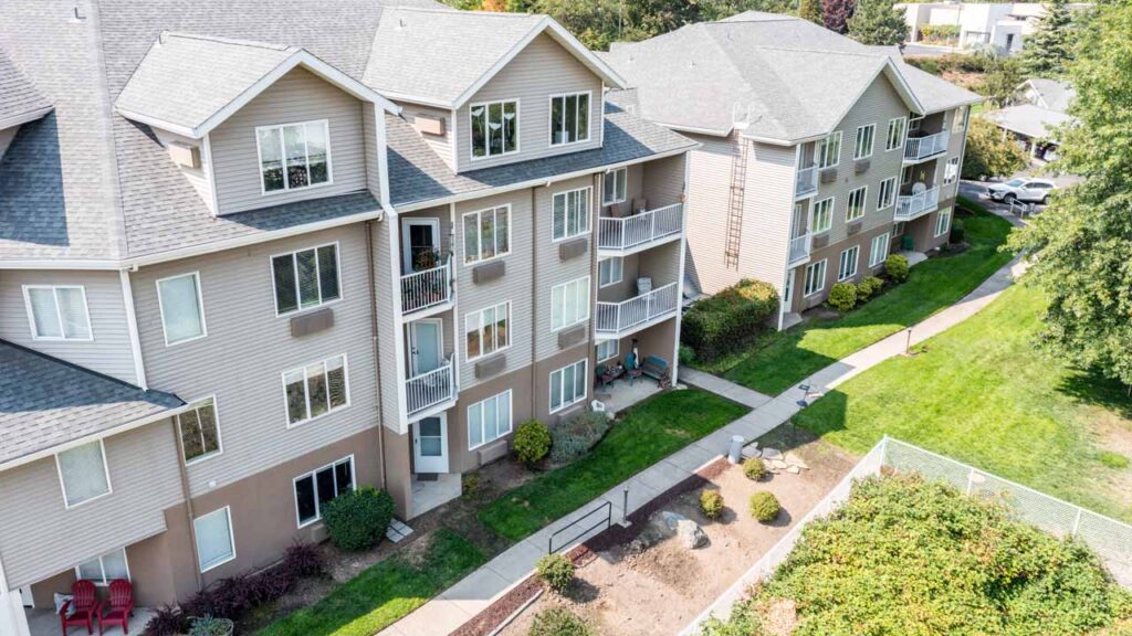 Aerial view of two beige apartment buildings with balconies, surrounded by green lawns, shrubs, and a sidewalk, on a sunny day.