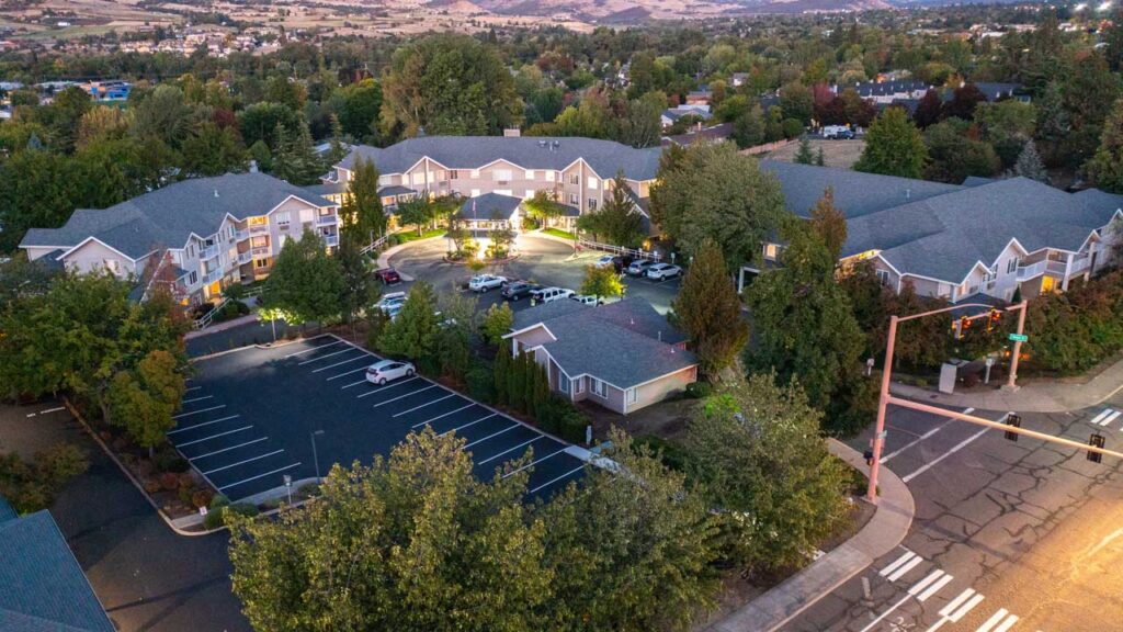 Aerial view of a large residential building with a central circular driveway and surrounding trees, parking lots, cars, and intersecting roads in a suburban neighborhood at dusk.