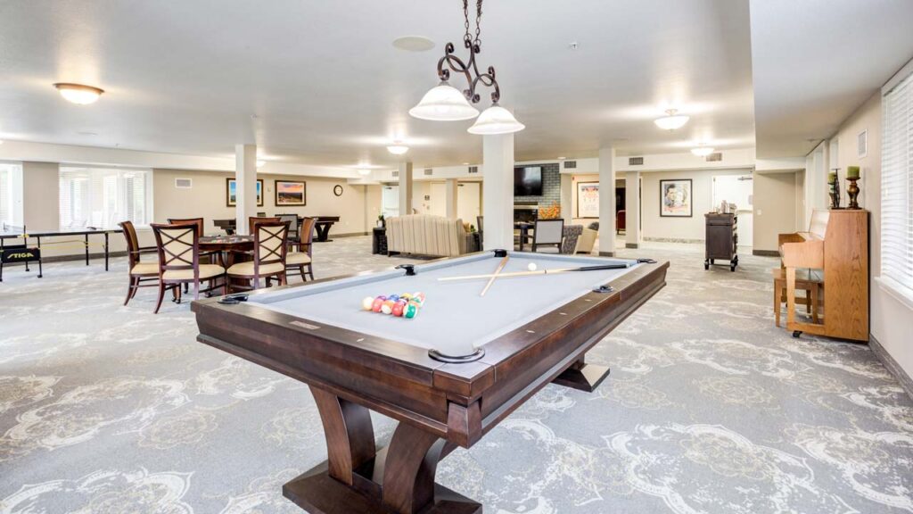 A spacious, well-lit game room with a pool table in the foreground, a table tennis setup, dining tables with chairs, a piano, and a seating area with a TV and fireplace in the background.