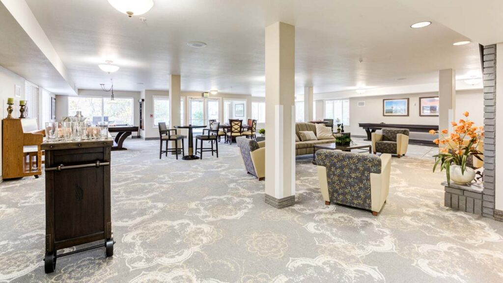 A spacious, well-lit lounge with patterned carpet, several seating areas, tables, pool tables, and large windows. Orange flowers in a vase and framed art decorate the room. The atmosphere is calm and inviting.
