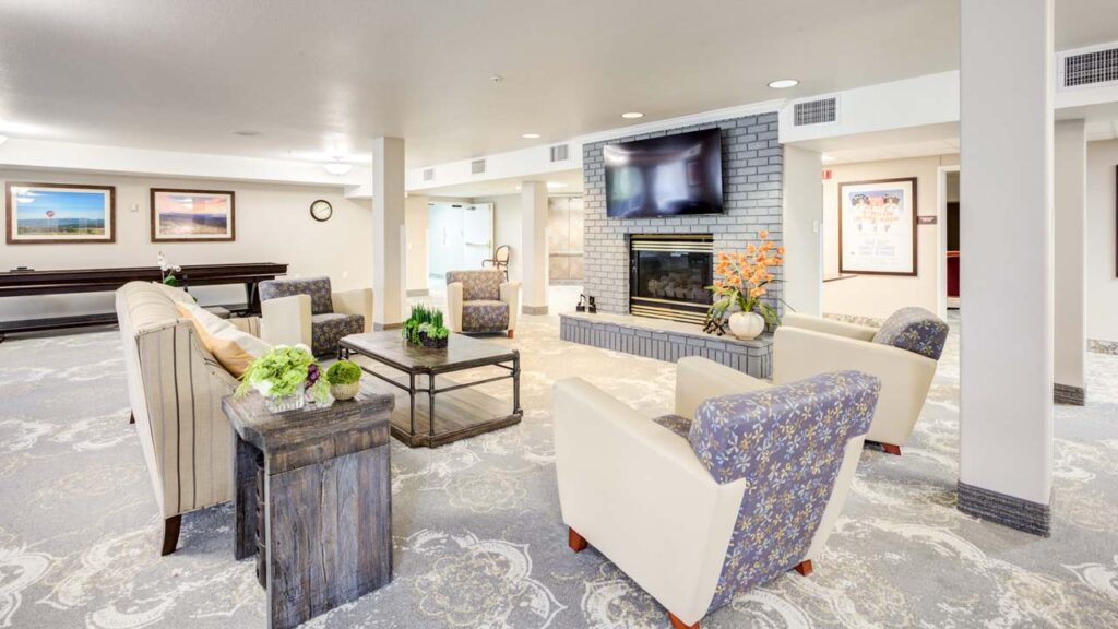A bright, spacious lounge with armchairs and a sofa arranged around a coffee table, a fireplace with a TV above it, decorative plants, and framed artwork on the walls.