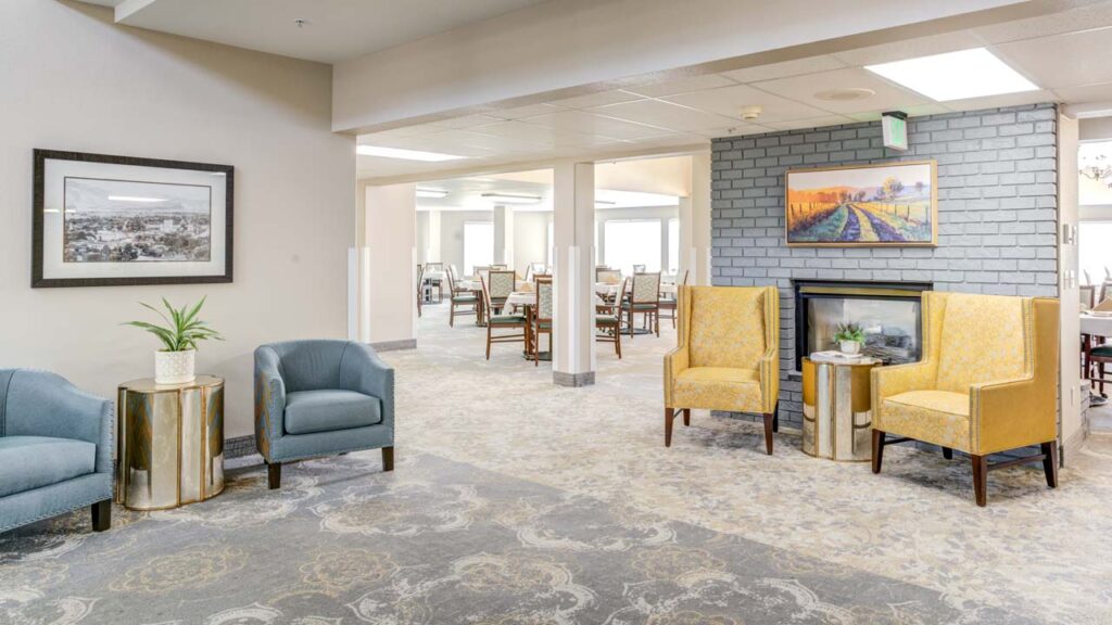 A bright, spacious lounge area with two blue armchairs, two yellow chairs by a fireplace, small tables with plants, framed artworks on the walls, and a dining area visible in the background.