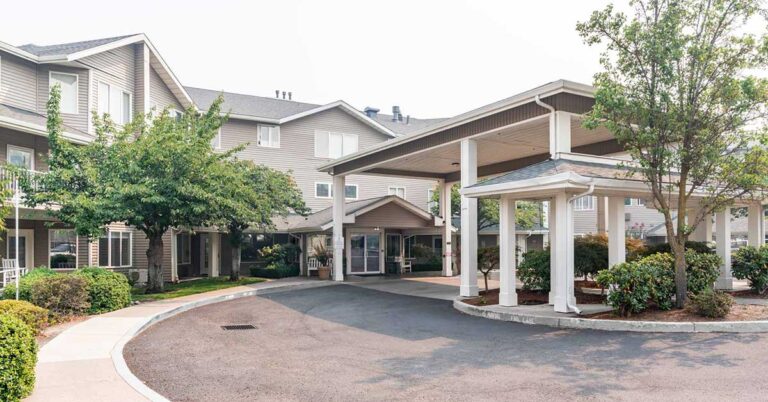 A modern assisted living facility with a covered entrance, surrounded by trees, shrubs, and well-maintained landscaping on a sunny day.