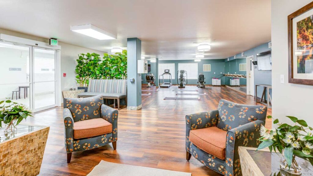 A bright, spacious room with wood flooring, two patterned armchairs, a small table with flowers, green plants, benches, and exercise equipment in the background. Large windows let in natural light.