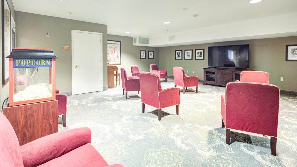 A small movie room with red cushioned chairs arranged in rows facing a large flat-screen TV. A popcorn machine stands in the corner, and framed pictures hang on the walls. The room has patterned carpet and soft lighting.