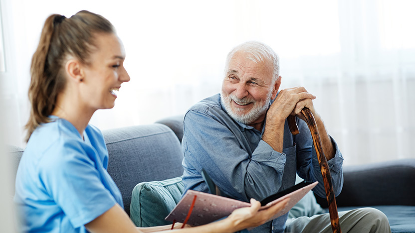 A young female caregiver in blue scrubs smiles while talking to an elderly man with a beard who is sitting on a couch, holding a cane and looking at her happily.