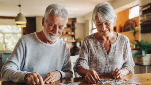 Two older adults sit at a table, smiling as they work together on a jigsaw puzzle in a bright, cozy room. They appear relaxed and content, enjoying each other's company and the shared activity.