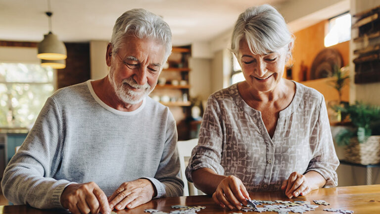 Two older adults sit at a table, smiling as they work together on a jigsaw puzzle in a bright, cozy room. They appear relaxed and content, enjoying each other's company and the shared activity.