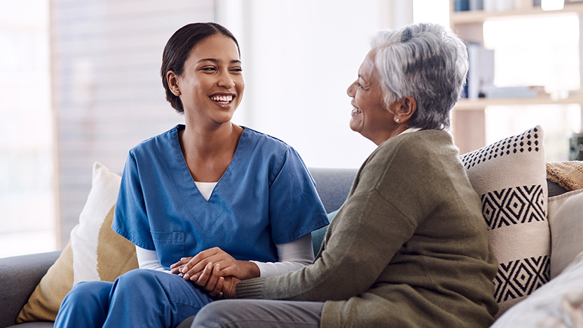 A young healthcare worker in blue scrubs sits on a couch, smiling and holding hands with an older woman with gray hair. They both appear happy and engaged in a warm, friendly conversation.