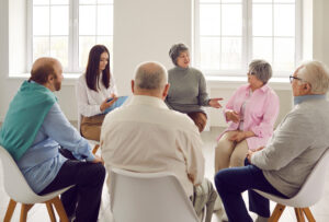 A group of older adults sit in a circle on chairs in a bright room, engaged in a discussion. One woman gestures while others listen. A younger woman takes notes on a clipboard.