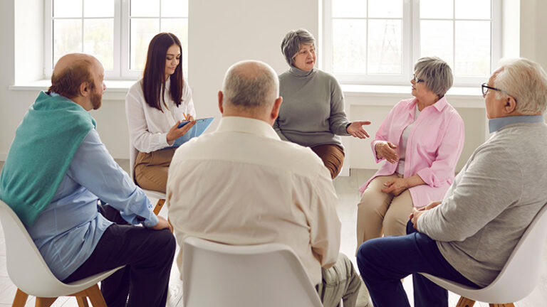 A group of older adults sits in a circle having a discussion, with one younger woman taking notes. In this bright, sunlit room, they participate in Memory Care Support Groups for shared understanding and connection.