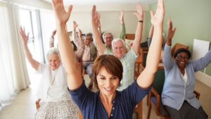 A group of seniors and a younger woman sit on chairs in a bright room, smiling and raising their arms enthusiastically during a group exercise session, often seen in Memory Care Support Groups.