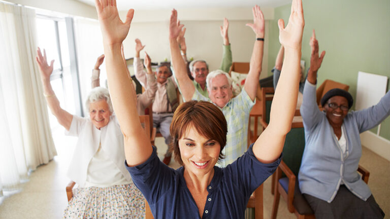 A group of seniors and a younger woman sit on chairs in a bright room, smiling and raising their arms enthusiastically during a group exercise session, often seen in Memory Care Support Groups.