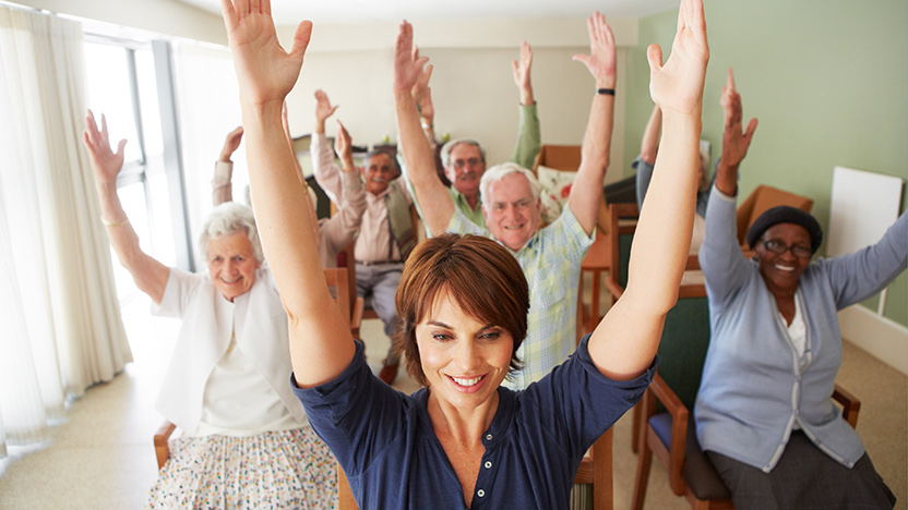A group of seniors and a younger woman sit on chairs in a bright room, smiling and raising their arms enthusiastically during a group exercise session, often seen in Memory Care Support Groups.