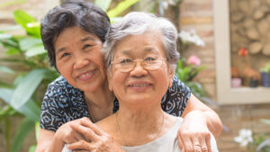 Two elderly women are smiling and embracing outdoors, radiating the joy of functional fitness for seniors. One woman, wearing glasses, sits in front while the other stands behind her with arms around her shoulders amid lush plants and a brick wall.