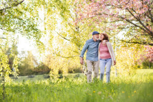 An older couple walks arm in arm through a sunny, blossoming park, soaking up sunshine for healthy aging as they enjoy a peaceful spring day amid green grass and trees with pink flowers.