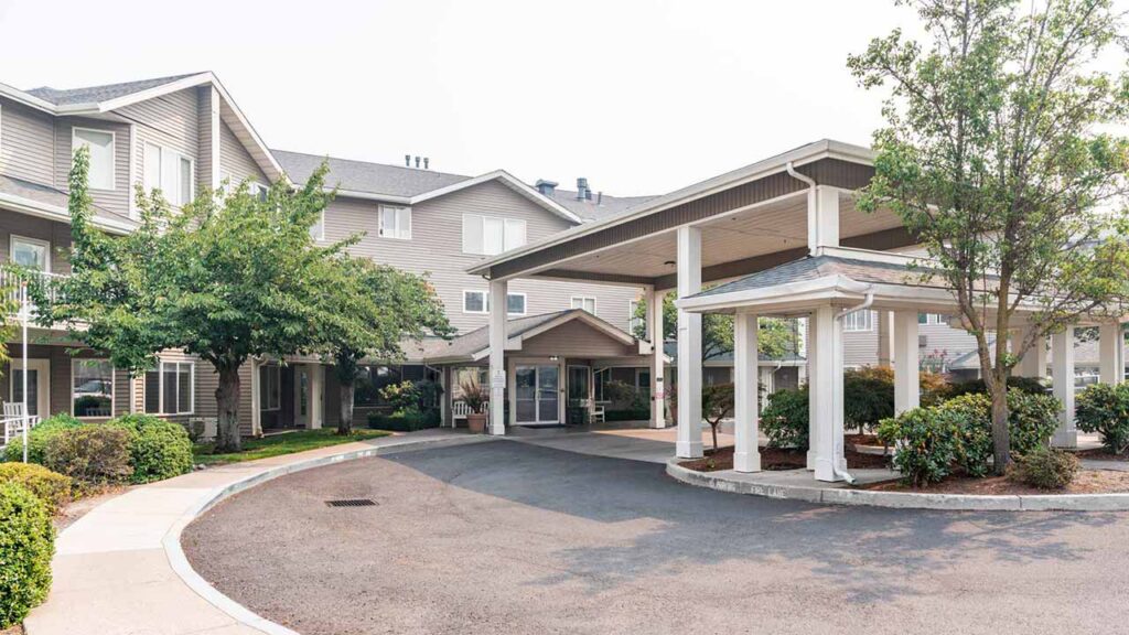 Exterior view of a multi-story residential building with a covered driveway entrance, surrounded by trees, shrubs, and neatly landscaped grounds on a clear day.