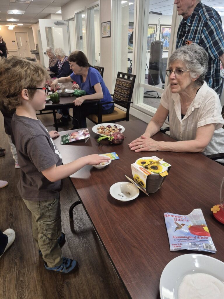 A young boy stands at a table handing seed packets to an elderly woman, highlighting senior learning as other seniors and children interact around tables in a bright, indoor community setting filled with gardening items.
