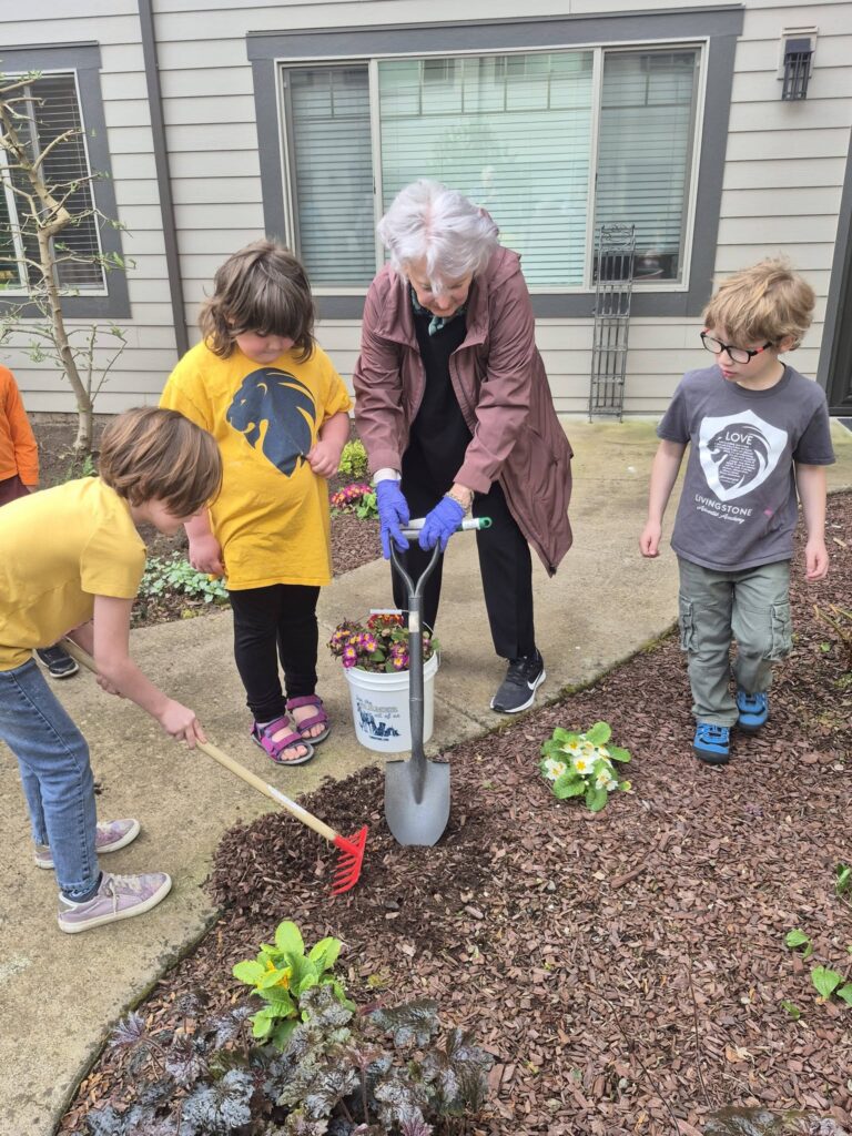 Four children and an older woman engage in senior learning while gardening outside a building. The woman holds a shovel and flower pot, guiding the children as they use garden tools, all focused on planting together.
