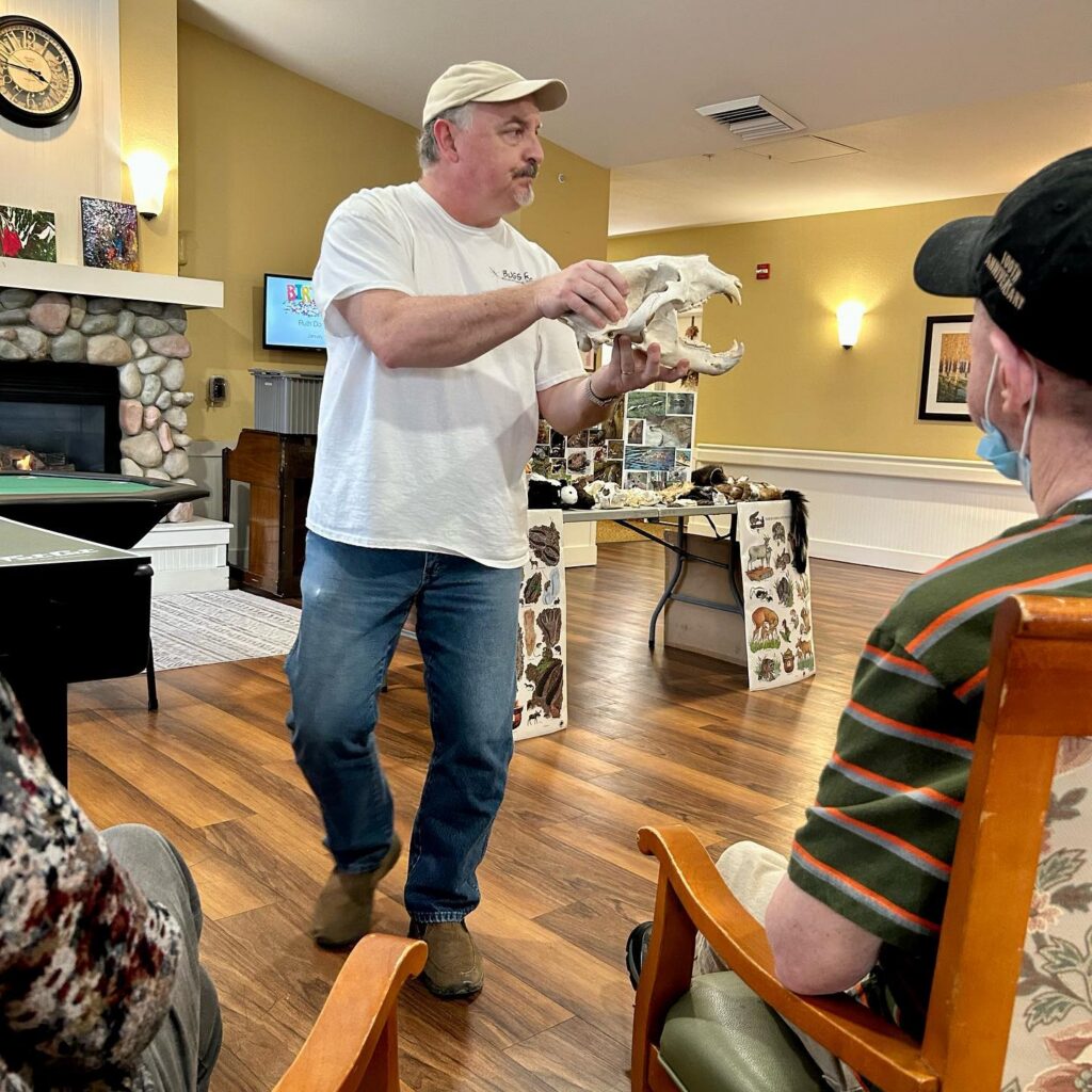 A man in a white shirt and cap holds up an animal skull while speaking to a seated audience, engaging in senior learning, in a room with wooden floors and a fireplace. A display table with animal artifacts is visible in the background.