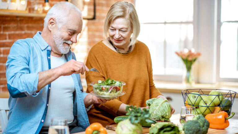 An older man and woman smile while preparing a fresh salad together in a bright kitchen, surrounded by superfoods for longevity like cabbage, broccoli, peppers, and apples.