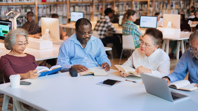 A group of four older adults engages in senior learning at a library table, reading books and using a laptop, with bookshelves and other people working in the background.