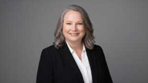 A woman with shoulder-length gray hair, dressed in a black blazer and white shirt, smiles warmly in front of a plain gray background, reflecting the confidence and vitality often seen in today’s senior living communities.