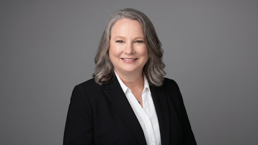 A woman with shoulder-length gray hair, dressed in a black blazer and white shirt, smiles warmly in front of a plain gray background, reflecting the confidence and vitality often seen in today’s senior living communities.
