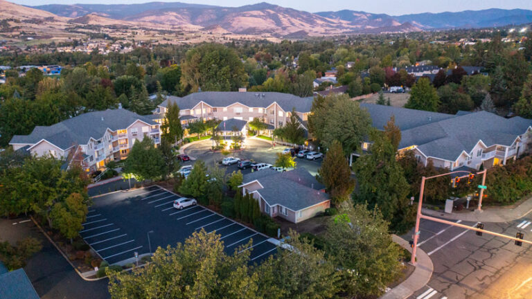 Aerial view of a spacious senior living residential complex surrounded by trees, with a well-lit circular driveway, parking areas, nearby intersection, and hills in the background.