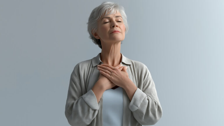 An elderly woman with short gray hair stands against a plain background, closing her eyes and resting both hands on her chest, embodying the calm and peaceful spirit often found in senior living.